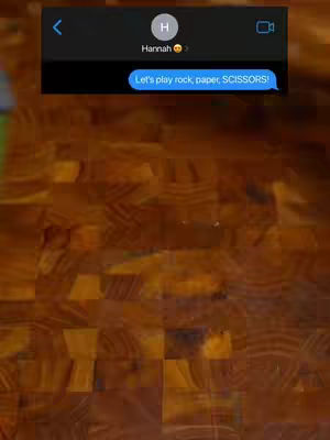 The cover image shows a screenshot of a text message conversation on a phone, with the text bubble stating, 'Let's play rock, paper, SCISSORS!' The background is a close-up of a wooden surface composed of small, tightly packed rectangular blocks of varying brown tones, suggesting a butcher block or similar surface.