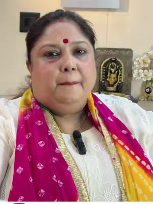 A woman with fair skin and dark hair gazes directly at the camera. She has a red bindi on her forehead. She wears a white shirt with a pink and yellow scarf draped around her shoulders. A gold chain with a black pendant hangs around her neck.
