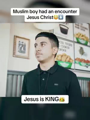 A man in a black polo shirt is centered in the frame with a slight upward gaze. Above him is a text overlay that reads “Muslim boy had an encounter Jesus Christ” with an emoji of a face with hearts for eyes followed by a downward arrow emoji. Below the man, another text overlay states “Jesus is KING” and includes a crown emoji. The background shows a restaurant interior with menu boards, food displays and what appears to be lights on the ceiling.