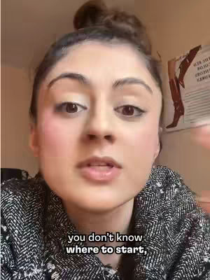 A close-up shot of a woman facing the camera, her hair pulled back in a bun. She has visible makeup and is wearing a patterned top. The background is blurred, but a section of a wall and what looks like a poster or artwork are visible. The bottom of the image includes superimposed text.