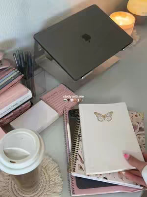 A top-down shot of a desk with pink and white themed study supplies. There's a laptop on a stand at the top, with a stack of books on the left, sticky notes and paper on the middle. A hand holding a notebook on the right. The background is blurred to a soft focus.
