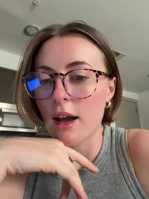 A woman with short brown hair and glasses looks towards the camera, her mouth slightly open. She wears a grey tank top, and a gold earring is visible. The background is a blurred indoor space.