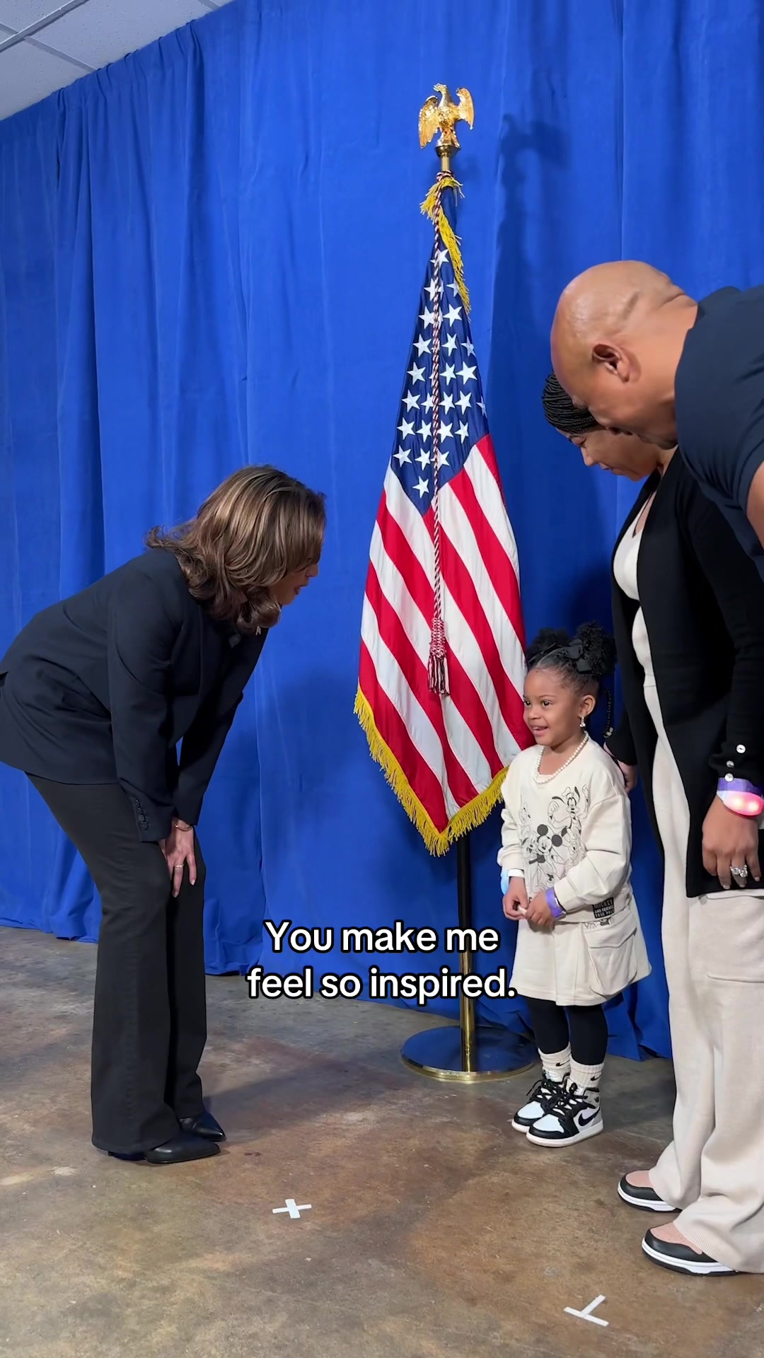The image shows Kamala Harris and two adults looking down at a young girl who is smiling, in a setting that appears to be some formal event. The background consists of a blue curtain behind the American flag. The American flag stands tall, with the stars and stripes clearly visible. The visual composition is balanced, with the figures on the right side of the frame and Harris on the left. The color palette is comprised of the flag's red, white, and blue, contrasted by the darker tones from the suits.
