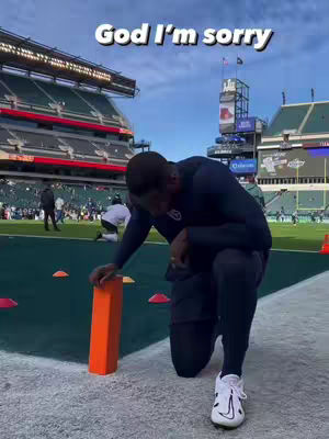 A person kneels on a football field, facing away from the camera. The person is dark-skinned, wearing a dark navy athletic outfit, and white and black athletic shoes. An orange marker is in front of the person. Background includes a stadium and blue sky.