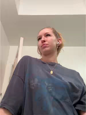 A young woman is looking up and to the side, with a slightly confused expression. She is wearing a gray t-shirt and a gold necklace. The background appears to be a white ceiling.