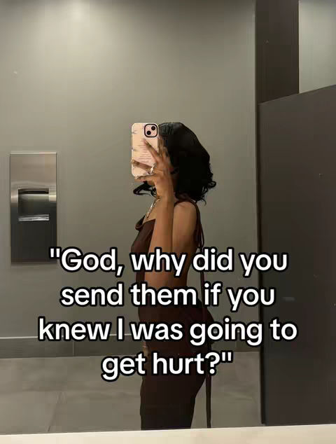 A woman is taking a selfie in a bathroom, holding the phone up to her face. Her dark hair is styled in loose waves. She is wearing a brown dress. The background is a mirror and a gray wall.
