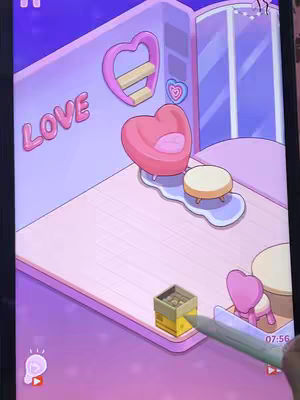 The cover image shows a digitally rendered room interior, viewed from a slightly high angle. The room has rounded walls painted in soft pink and purple hues. Inside, there is a heart-shaped chair, a small circular table, a heart-shaped shelving unit, and a rug that resembles a puddle. Additionally, a hand holding a digital pen is partially visible, interacting with the objects on the screen. The overall style is cute and whimsical, and evokes a sense of a game or app interface.