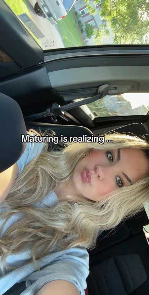 The cover image is a selfie taken inside a vehicle with the creator facing towards the camera. The foreground features the creator with long blonde hair, light makeup, and a slight smirk. The background reveals a view of a residential area through the car's windows, with green trees and part of a building visible. The lighting appears natural, and the overall composition is an intimate shot.