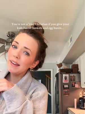 A woman with her hair up in a messy bun is in the foreground, smiling softly. She's wearing a gray cardigan. Behind her, a kitchen is visible, with a refrigerator covered in photos and a cabinet. The lighting is bright, suggesting natural daylight.
