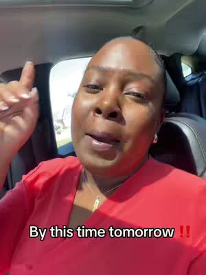 A woman with dark skin and short hair is seated in a car, angled slightly towards the camera. She wears a red top and a gold necklace. The background is blurred, showing the car's interior and the outside environment, which appears to be daylight. Text is overlaid at the bottom of the frame.