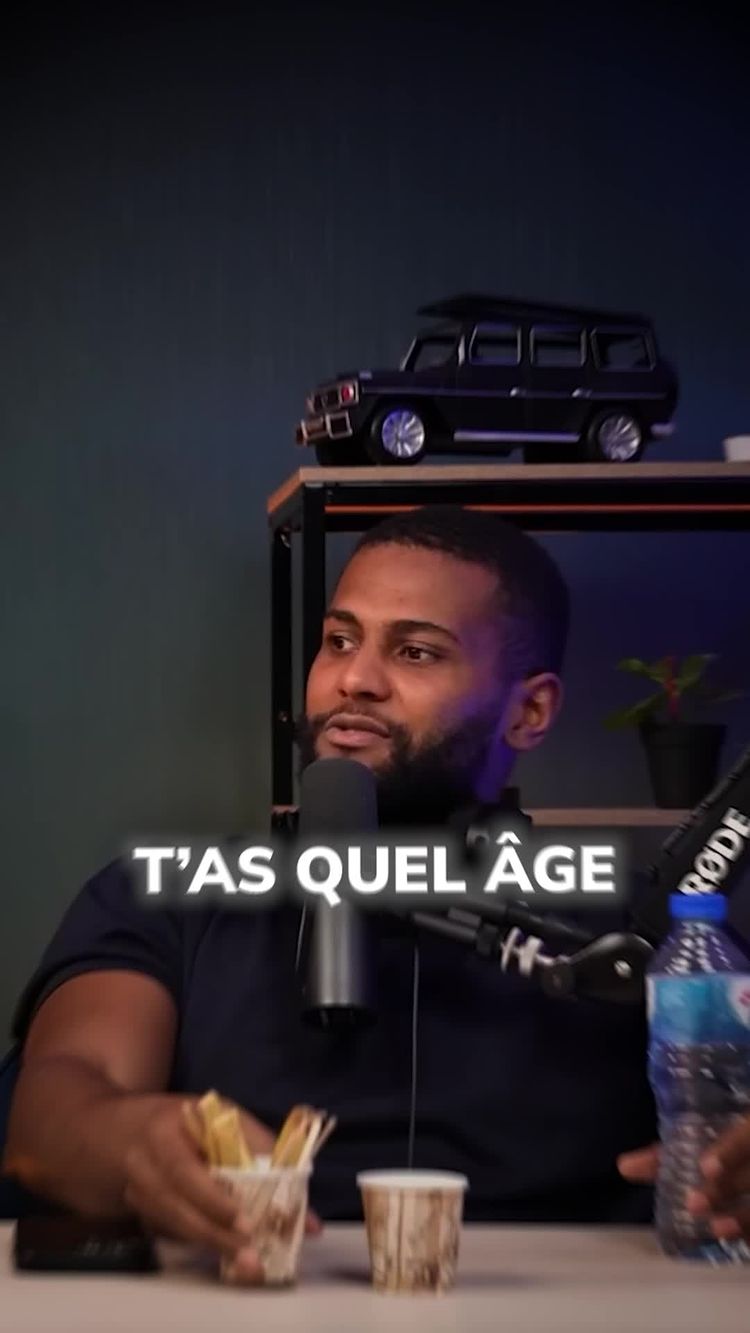 The image shows a man in a dark blue shirt speaking into a microphone. The text overlay that says T'AS QUEL ÂGE is placed in the front left. Behind him is a small shelf with a toy car on top of it. There's also a bottle of water and a drink next to them. The background is a dark gray color.