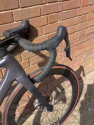 The image showcases a partial view of a black road bike, set against a backdrop of a reddish-brown brick wall. The bike's handlebars, wrapped in black tape, curve towards the viewer, with a portion of the front wheel and frame visible. The lighting is bright, highlighting the textures of the materials. The bike's tire has a red rim. The image composition favors a close-up angle, focusing on the details.