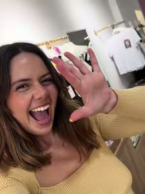 A selfie taken from a slightly low angle shows a smiling woman with her mouth wide open, as if laughing exuberantly. She has brown hair and is wearing a yellow sweater. Her right hand is raised towards the camera, with her fingers spread. The background is blurred, but suggests a retail or shop environment.
