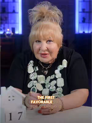 A woman with blonde hair in a bun is centered, wearing a black shirt with light decorations. A decorative necklace and bracelet are visible. The bottom of the image shows a white house with the number 17 on it, and text overlaid in the middle with gold text.