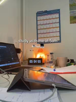 The image shows a desk setup for studying. A laptop is open on the left, and a tablet with a cover is propped up in front of it. A digital clock displays the time, "05 04 05". There's a mug, a water bottle, and some papers on the desk. A calendar hangs on the wall in the background.