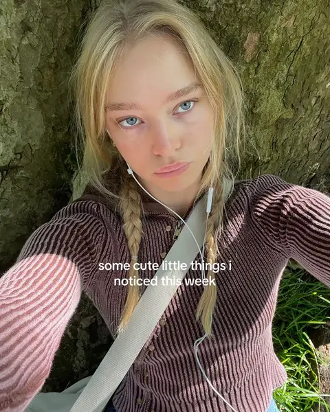 A selfie taken from a slightly above angle shows a young woman with blonde hair, styled in two braids, looking at the camera. Her skin is fair, and she has blue eyes. She wears a light brown sweater and neutral-colored headphones. The background is a blurry view of a tree trunk and foliage. Text overlay at the bottom.