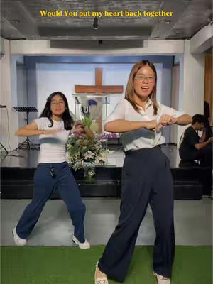 Two young women are dancing in a church. One is wearing a white top and blue pants, the other is wearing a white top and black pants.