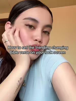 A young woman is seen in a selfie-style shot with her face close to the camera. She has dark hair, fair skin with visible makeup, and is wearing a light blue shirt. She's touching her cheek with one hand, and the background is a blurred, warm-toned interior. Text overlayed on the image provides additional context.