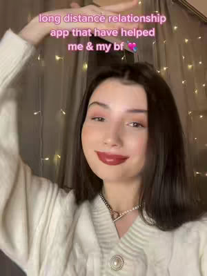 A woman with dark hair and a white sweater smiles at the camera. Text overlayed on the image includes the words about long distance relationships. The background is softly lit with fairy lights, creating a warm atmosphere.