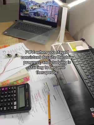 A desk setup with a laptop displaying a cityscape on its screen, a calculator, and papers strewn across the surface. A small desk lamp is positioned to the side. Additional items include a pencil, headphones, and sticky notes. The background is blurred.