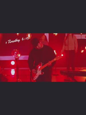 A live performance stage bathed in red light. A man playing an electric guitar is the central figure, with other performers in the background. Text overlays are present.