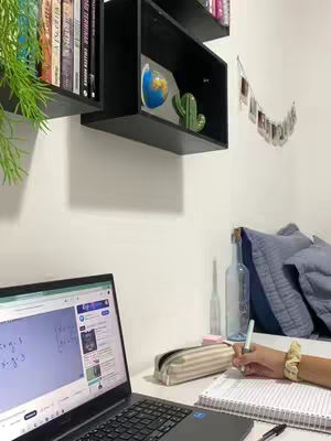 The image shows a well-lit study space with a laptop displaying math equations, a notebook, and a hand writing. Bookshelves, a shelf with a globe and cactus, and a photo display add character. The composition is clean, highlighting the study setup.