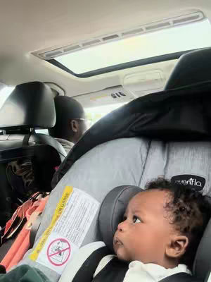 A car interior shot with a baby in a car seat. The baby is looking to the right, with a seat belt fastened. Another person is visible in the back seats and a sky roof above.