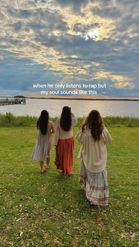 Three women are seen from behind, walking away from the camera across a grassy area towards a body of water. The sky is overcast with clouds, and there's a pier in the distance. Text overlayed on the image reads "when he only listens to rap but my soul sounds like this".