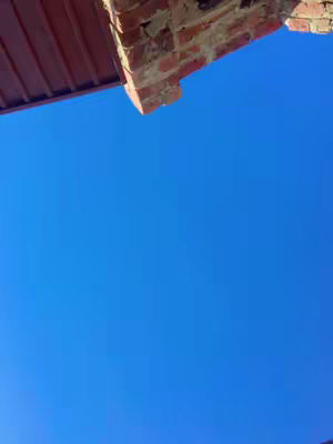 An upward shot capturing a clear blue sky as the primary backdrop, with a portion of a building's structure in the upper frame. A section of a reddish-brown roof or awning extends horizontally across the top left, transitioning to a brick wall. The bricks vary in color from light to dark red and brown, with visible mortar lines. The overall composition focuses on simplicity and natural aesthetics, employing complementary colors.