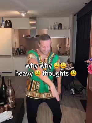 A man dances energetically in a kitchen setting, wearing a colorful, patterned shirt and black pants. The background includes kitchen cabinets, appliances, and a glimpse of the living area. Text overlays highlight the emotional tone with the words "why, why heavy thoughts" and sad face emojis. The overall composition is a medium shot with a focus on the man's dancing expression.
