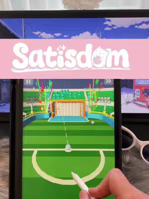 The cover image shows a person holding a tablet displaying a mobile game. The game interface shows a soccer field with colorful graphics, bright lights, and a cartoon-style aesthetic. The person's hand, holding a stylus, is visible in the foreground, positioned towards the bottom, suggesting interaction with the game. There's a banner at the top with the game's title in playful font overlayed on a pink background. In the background, there are two additional screens displaying related content.