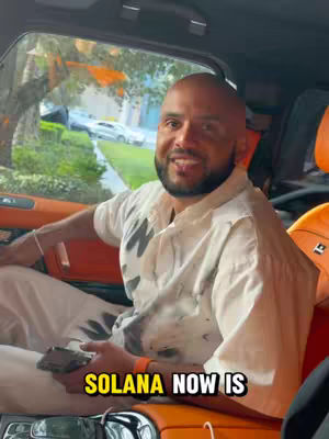 A man with a partially bald head and a beard is seated inside a car. The interior appears to be luxurious with orange and black leather. The man is wearing a white shirt with a print on the front. The text overlay says SOLANA NOW IS.