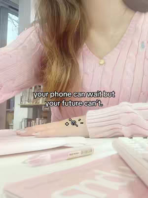 A close-up shot of a person sitting at a desk, wearing a pink sweater and working on a project related to studying. Text overlayed reads, 'your phone can wait but your future can't.' A pink highlighter pen is visible on the desk.