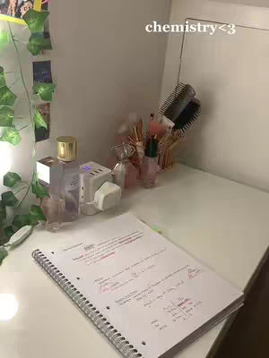 A well-lit, slightly angled shot of a desk with study materials. A notebook with notes occupies the foreground, and various cosmetic items and greenery sit on the desk. The photo is generally bright and airy.