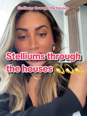 A woman with long blonde hair is centered in the frame, looking towards the camera. She is wearing a black top and a delicate necklace. Text overlays in white and pink read "Stelliums through the houses". The image is well-lit, with the woman's face being the focus, set against a simple indoor backdrop.