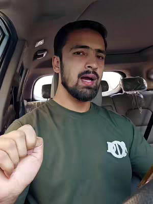 A man is seated in the driver's seat of a car, facing the camera with an open mouth expression. The background shows the interior of the car and an outdoors setting. His left hand is raised in a fist, with a logo on his shirt. The lighting appears natural.
