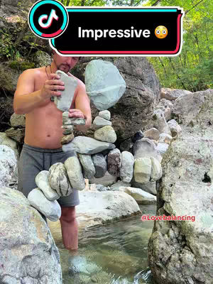A man is standing in a river, balancing rocks. He is shirtless, wearing gray shorts, and is focused on the arrangement of stones. A hook in a speech bubble is placed in the upper right with the text 'Impressive'. The photo is set in a natural, outdoor environment with large rocks and green foliage; the water is clear.