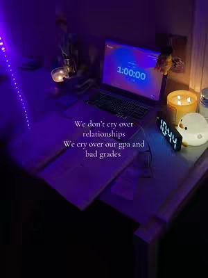The image shows a dimly lit desk with study materials and decorations. A laptop displays a timer, and text overlays convey a relatable struggle with academics.