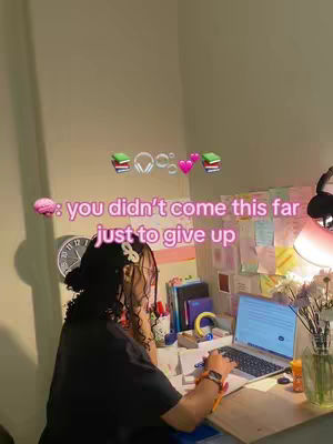 A person is sitting at a desk, working on a laptop. The person is seen from the back, wearing a black shirt, hair in a bun, and a hair clip. The desk is cluttered with study materials. There is a clock on the wall. Motivational text is overlaid on the image.