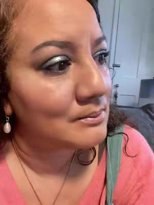 A close-up shot of a woman's face looking slightly to the side. She has brown eyes with silver eye shadow, dark eyebrows, and glossy lips. She wears a pink top and a gold necklace with a pendant. The background is blurred, but appears to be an indoor setting.
