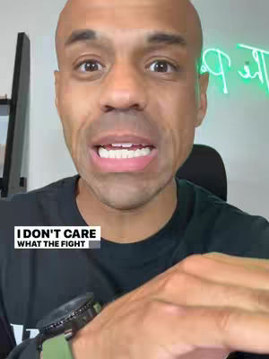 A bald man with light skin looks intensely at the camera, mouth slightly open. He wears a black t-shirt and a smartwatch, with a background showing a computer monitor and a neon sign. A text box overlay at the bottom left reads, “I DON’T CARE WHAT THE FIGHT”.