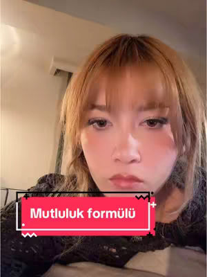 A selfie-style shot featuring a young woman with light skin and long, light brown hair which is parted on the side; the hair is slightly wavy and falls to her shoulders. She has a subtle makeup look with blushed cheeks and a nude lip. A red rectangular text box overlays the front of her chest, in which white text reads "Mutluluk formülü," translated as "Happiness Formula". The background is softly lit with warm tones, suggesting an indoor setting.