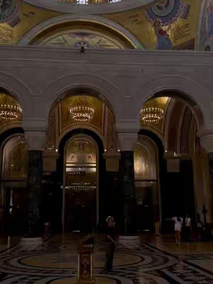 An interior shot of a grand church or cathedral featuring arched ceilings, ornate chandeliers, and a patterned floor; a person stands near the center in front of a podium or altar.