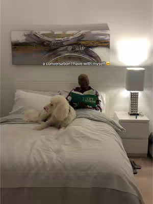 A person is sitting on a bed with a white dog, with the person reading a book. An image with an abstract painting is on the wall behind.