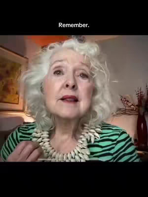 An older woman with light gray, curly hair is centered in the frame, looking slightly up and to the left. She has fair skin, and her lips are slightly parted. She wears a green and black striped top and a white beaded necklace. The background is blurred, with hints of artwork and possibly a red vase visible.