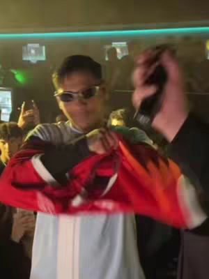 A person wearing sunglasses and a white shirt with red and black sleeves is holding a red jersey in front of a crowd. The person is in focus, with others blurred in the background. The setting seems to be indoors with lighting.