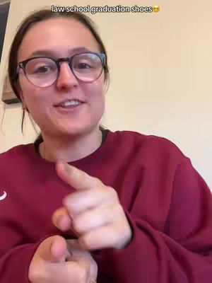 A woman with glasses is pointing at the camera with both hands. She is wearing a maroon sweater. The background is a simple, lightly colored wall.