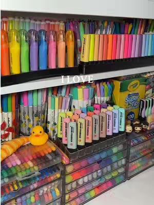 A well-organized display of various colorful stationery items, including highlighters, pens, pencils, and markers, arranged neatly on shelves and in clear drawers. The overall composition is bright and visually appealing, with a focus on the variety of colors.