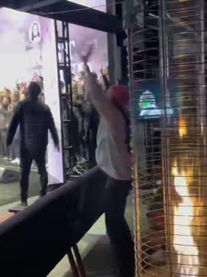 A scene at an outdoor worship event during night time with a person in a white shirt standing with their arms raised in worship, surrounded by other people, with staging elements on the left, and an outdoor heater with a visible flame on the right.