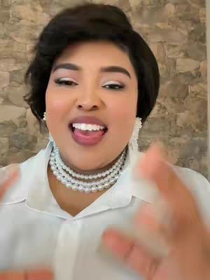 A woman with dark hair and makeup looks directly at the camera, mouth open in a smile, with her hands raised near her face. She wears a white shirt and pearl necklace. The background is a textured, light-brown wall.